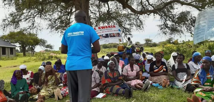 A group of people sitting under a tree outside listen to someone talking