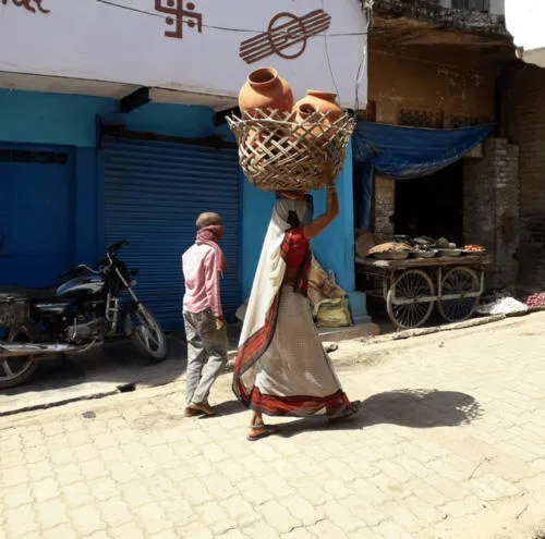 People walking up the street, one of which is carrying a basket of pots on their head