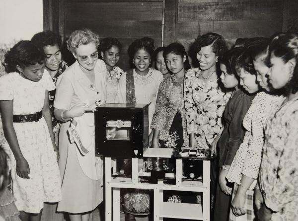 A white woman showing group of Malay women how to cook on a kerosene stove