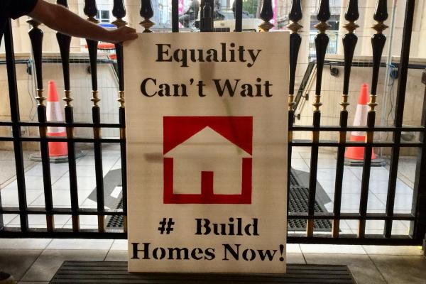 Person holding a sign that says 'Equality Can't Wait #Build Homes Now!'